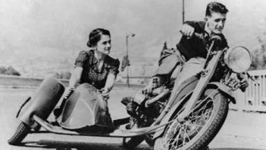 Man riding a motorcycle while a woman in the sidecar looks on affectionately.